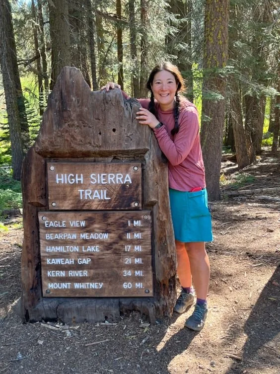 A hiker standing by the trailhead sign for the High Sierra Trail at Crescent Meadow, surrounded by giant sequoia trees.