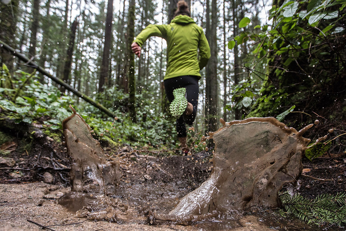 A woman trail runs through a wet, green forest and splashes through puddles.
