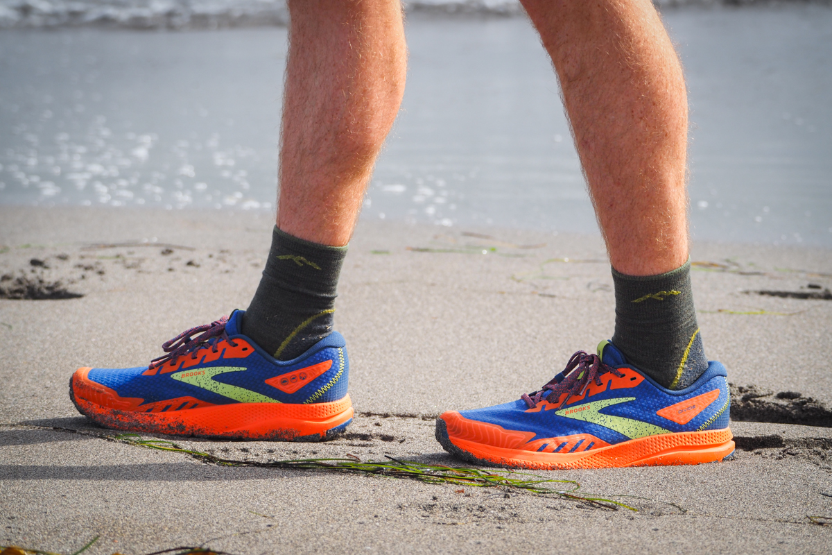 A runner wears Brooks Divide shoes while walking along a sandy beachfront terrain.