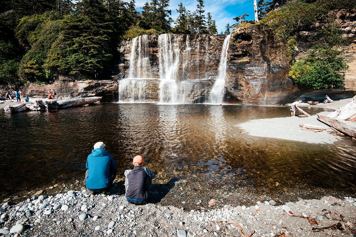 Two hikers sitting on a rocky beach admiring a tall waterfall.