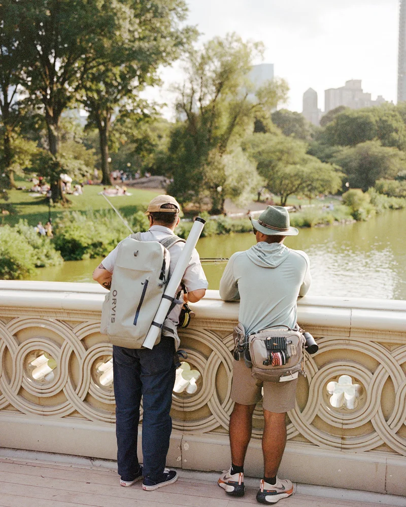 A fly fisherman standing near a historic stone bridge over a Central Park lake.