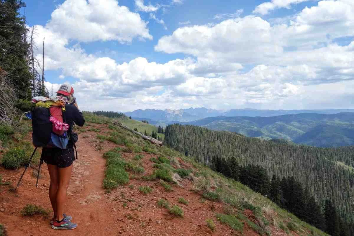 A backpacker standing on the trail overlooking a valley with mountain resort infrastructure.