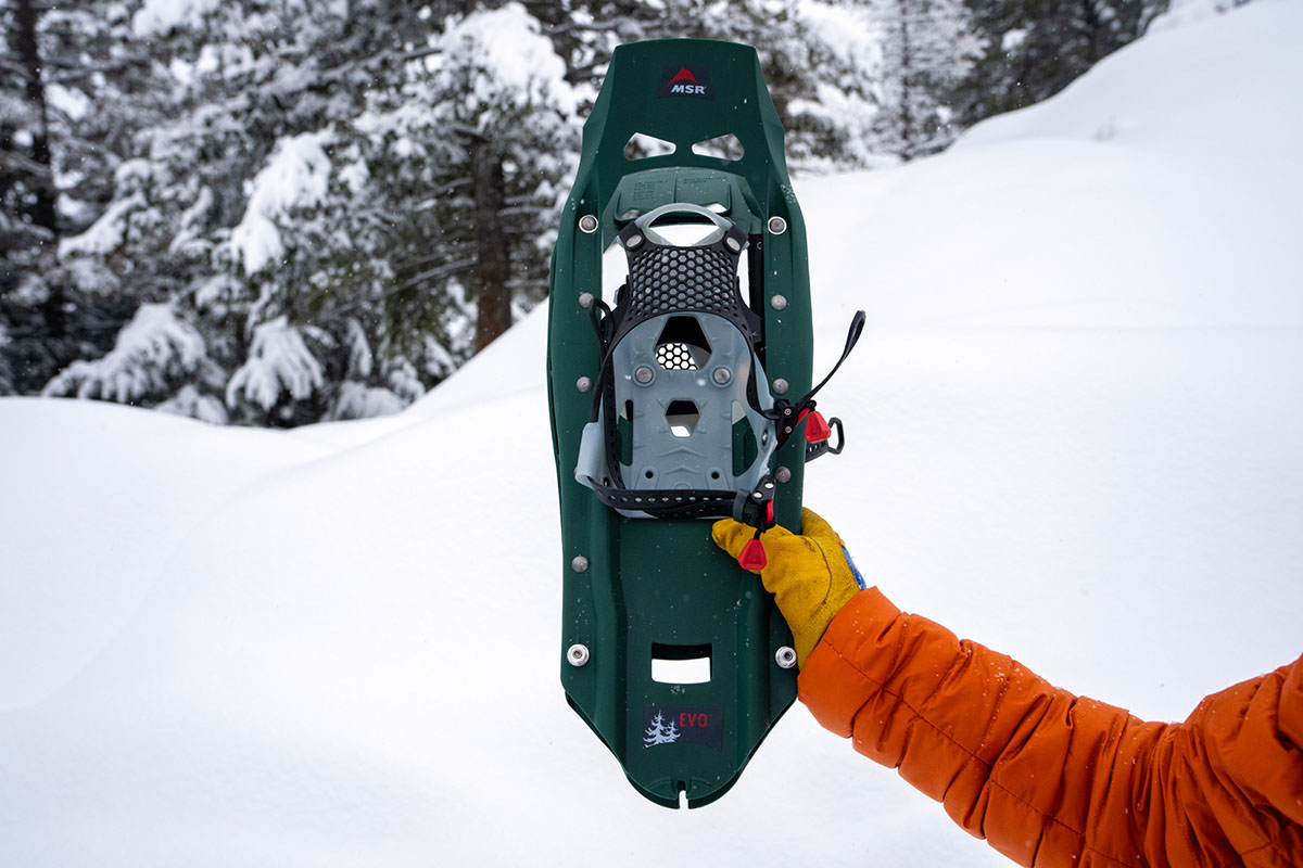 A hand holding a green MSR Evo Trail snowshoe in a snowy woods setting.
