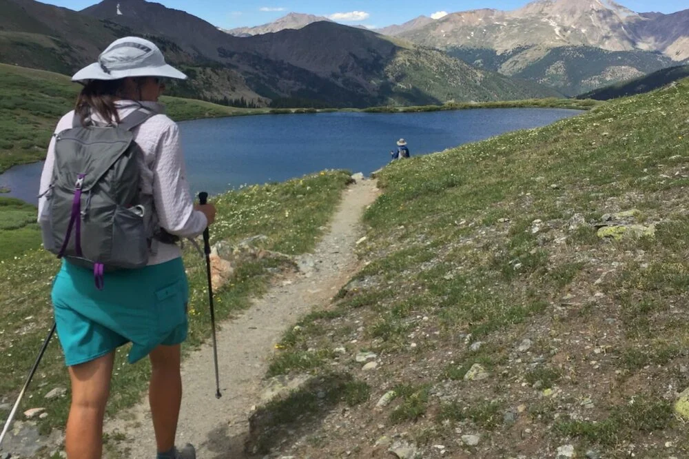 A hiker using trekking poles on a mountain trail in Colorado with a scenic mountain backdrop.