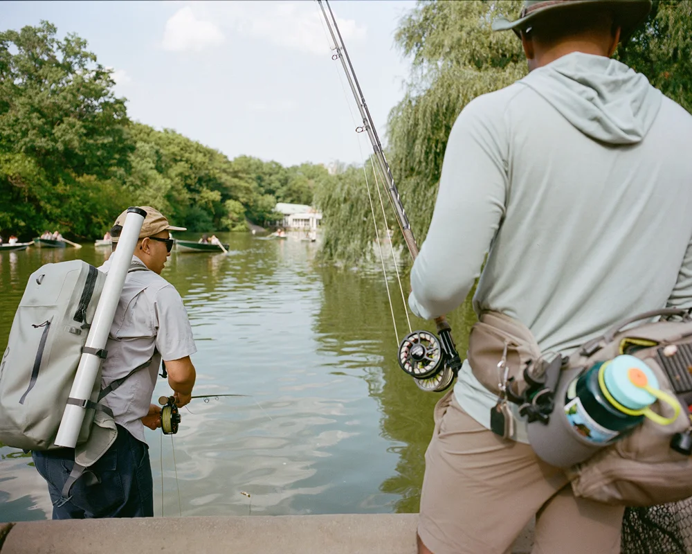 An angler fishing in Central Park's waters with the iconic Plaza Hotel and city skyscrapers looming behind.