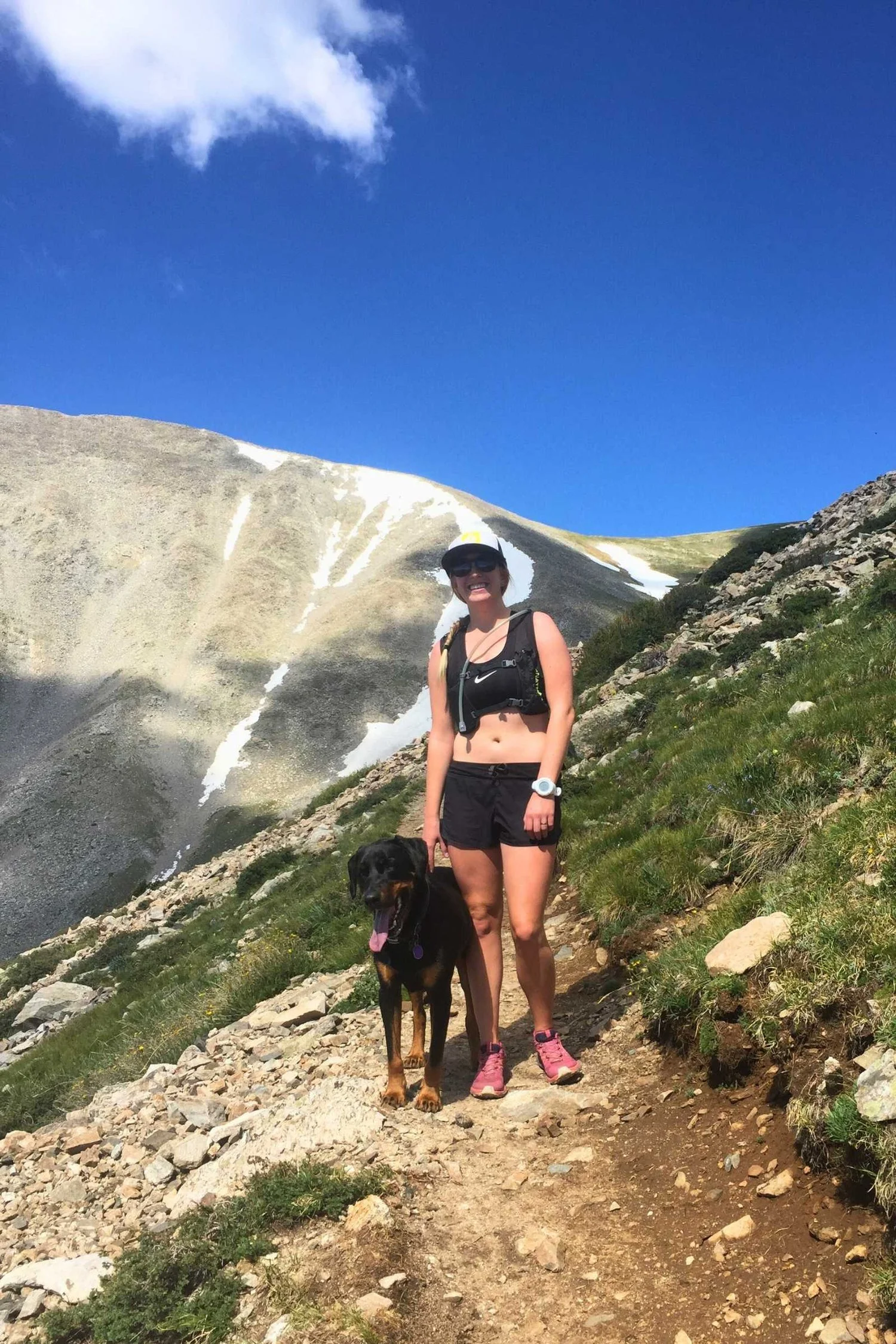 A woman hiking uphill on a sun-drenched trail wearing a black sports bra and carrying a daypack.