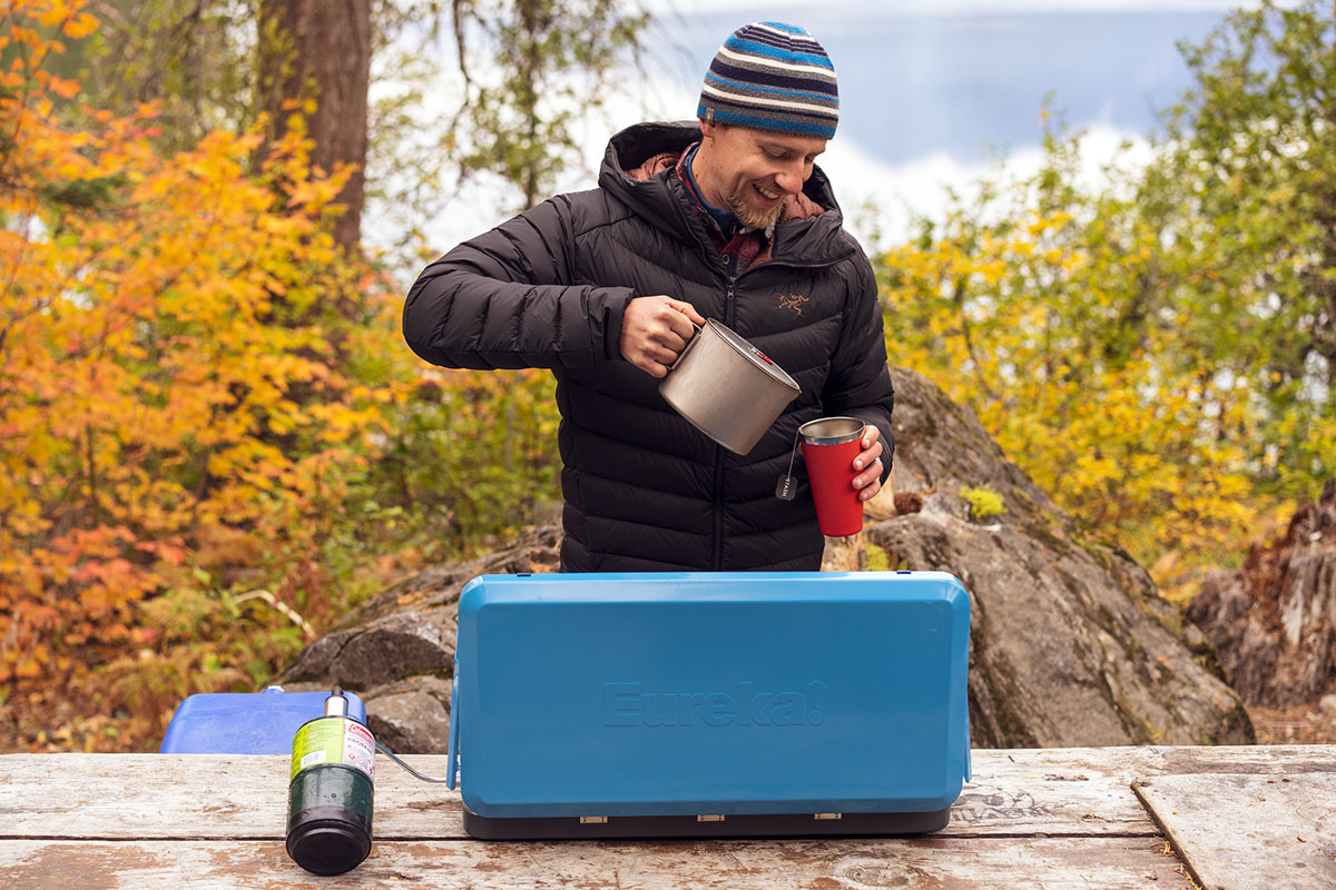 A person cooking breakfast on a blue Eureka Ignite Plus 2-Burner Camp Stove outdoors.