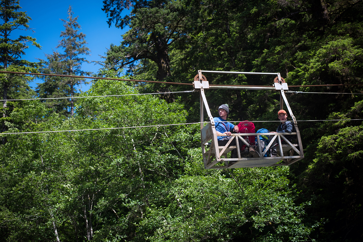 Two hikers sitting in a small, manual wooden cable car suspended over a forest gap.