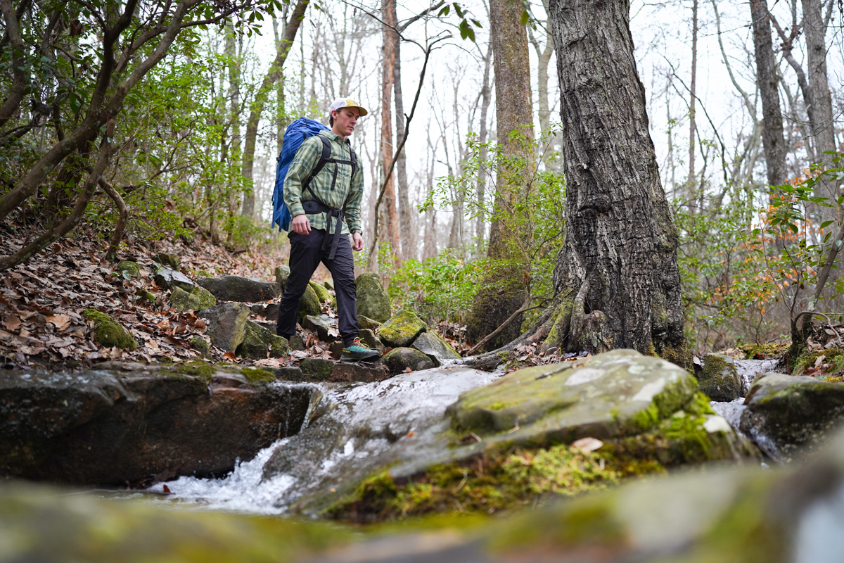 A hiker in a button-up hiking shirt walking near a creek, demonstrating ventilation options.
