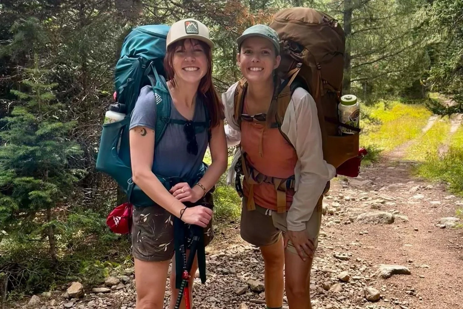 Two female hikers standing together on a trail, showcasing how hiking-specific bras fit under backpack straps.