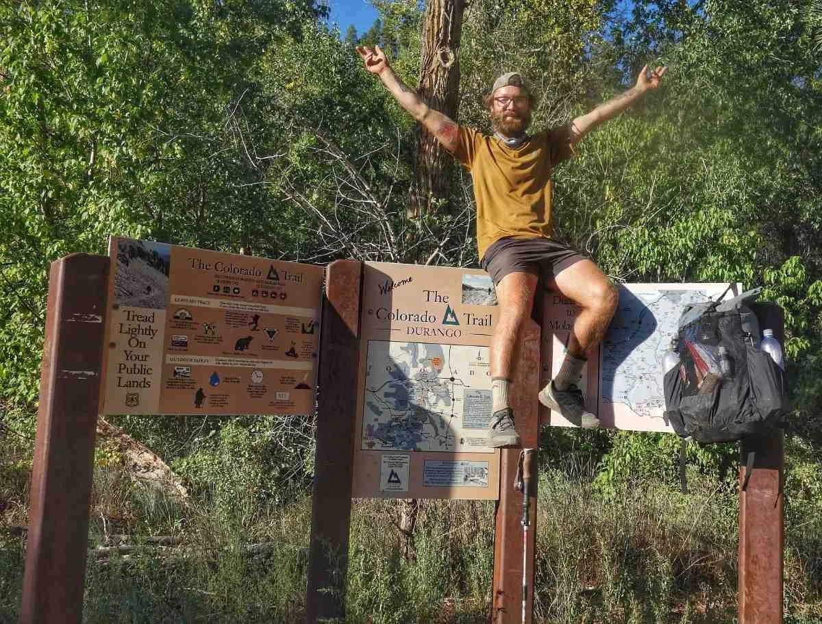 A happy hiker posing next to the official Colorado Trail Southern Terminus sign in Durango.