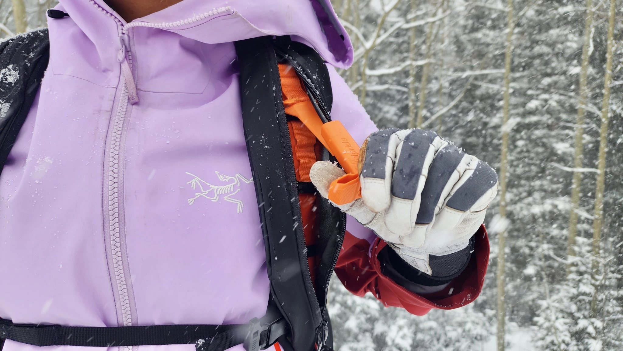A close-up of a skier's hand reaching for the deployment trigger on the shoulder strap of an avalanche pack.