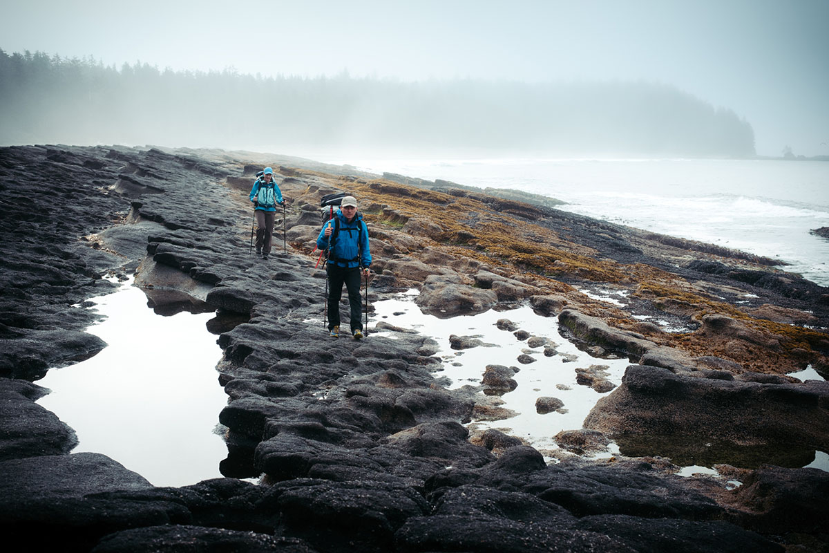 Two hikers walking along a foggy, rocky coastline on a dark day.